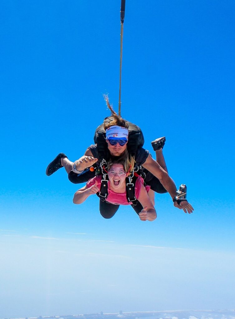 Two people tandem skydiving against a clear blue sky. The instructor, wearing a blue helmet and sunglasses, is positioned behind and above the person in a pink shirt, who smiles with arms outstretched while descending.