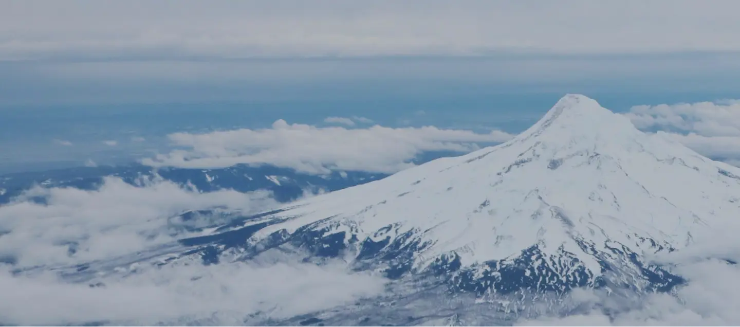 Aerial view of a snow-covered mountain peak surrounded by clouds. The mountain's surface is marked with dark rocky patches beneath the snow, creating a striking contrast. The sky above is overcast, blending with the clouds around the peak.