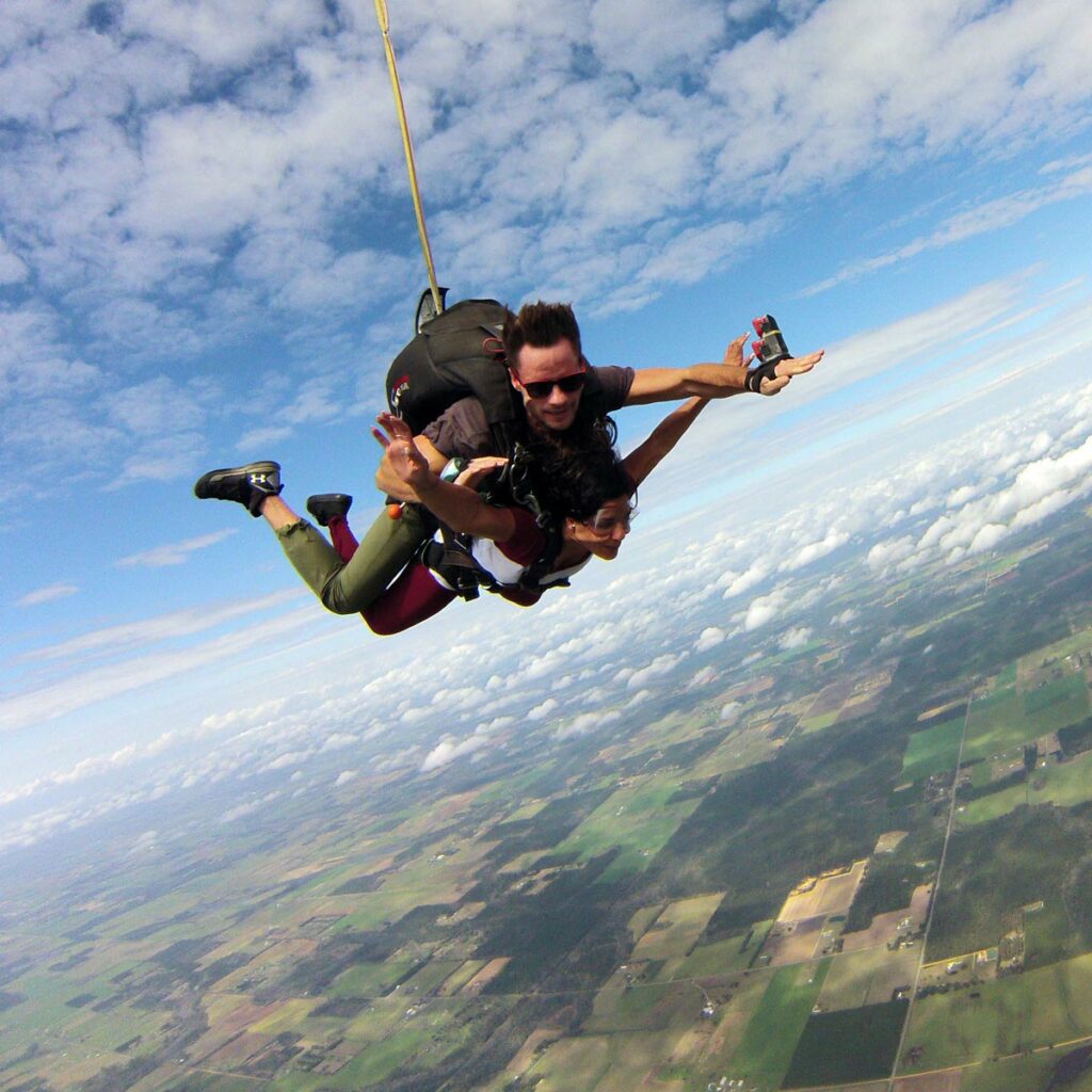 Two people tandem skydiving, freefalling through a partly cloudy sky with one person wearing dark sunglasses and holding an action camera. The landscape below features green fields and patches of farmland.
