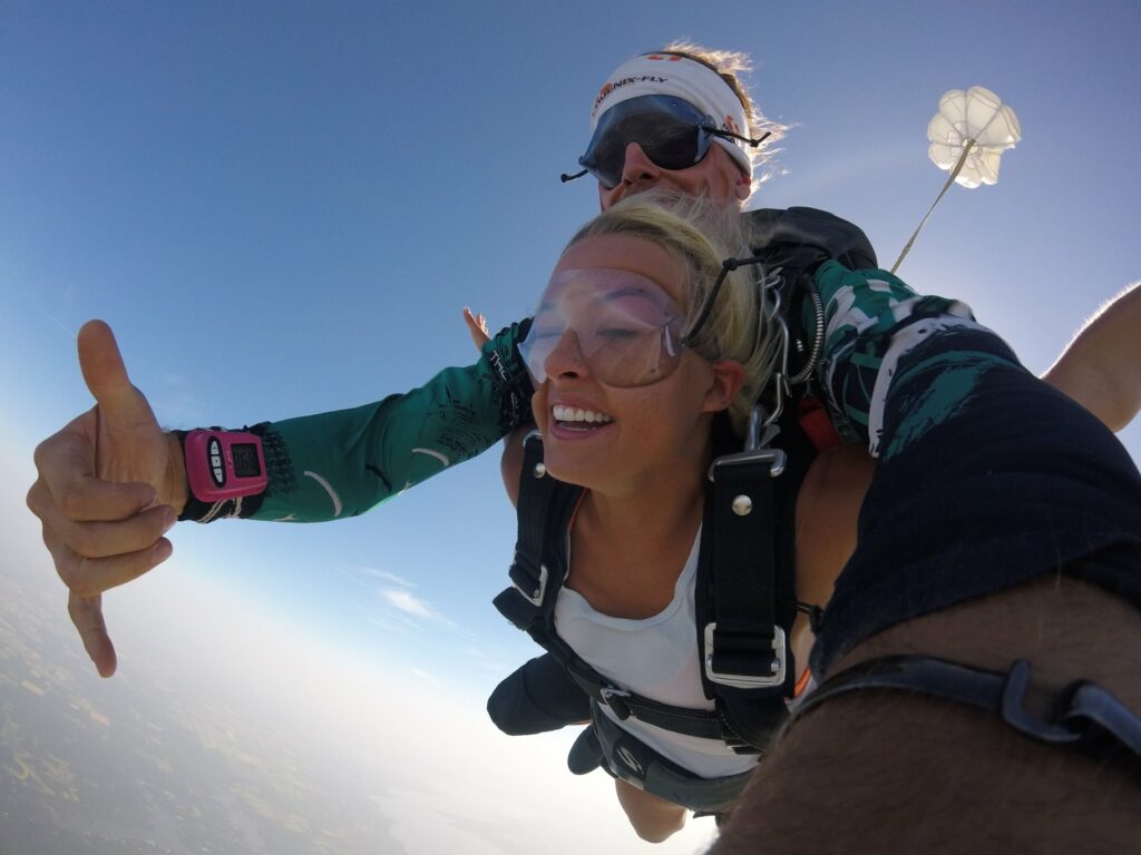 Two people tandem skydiving against a clear blue sky. The person in front smiles broadly, wearing goggles and a white tank top. The skydiving instructor is behind, wearing sunglasses and a headband. A parachute trails above them.