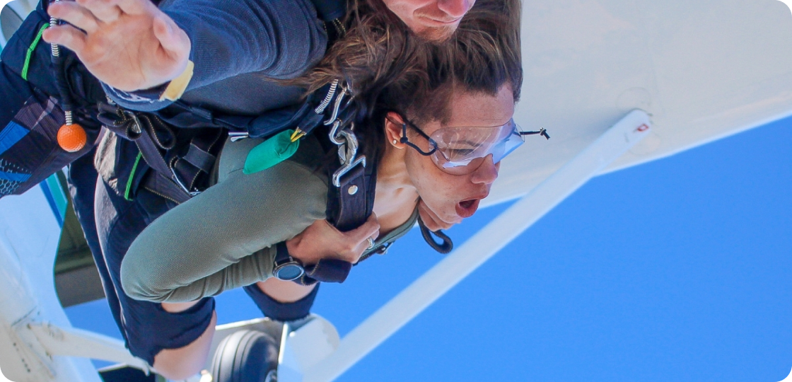 A woman tandem skydiving with an instructor, both wearing harnesses and goggles, as they exit an airplane. The woman appears excited and thrilled. The sky is clear and blue in the background.