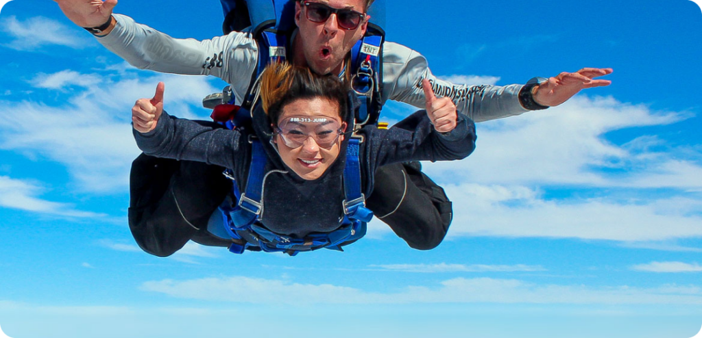 A person tandem skydiving with an instructor over a blue sky. They are giving thumbs-up while wearing goggles and harnesses. The instructor is behind and slightly above, both looking excited.
