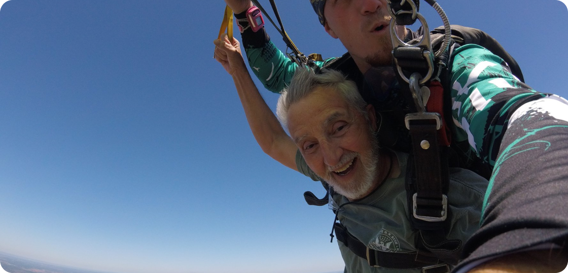 Two people tandem skydiving; an older man is in front, smiling joyfully, with a younger man at his back controlling the descent. They are high above the ground with a clear blue sky in the background.