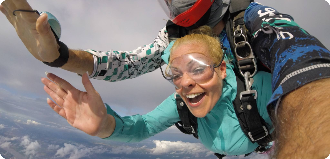 A person with a wide smile is skydiving, tandem with an instructor. They are both wearing goggles, and the person in front has blonde hair and a turquoise jumpsuit. The background shows a cloudy sky and horizon.
