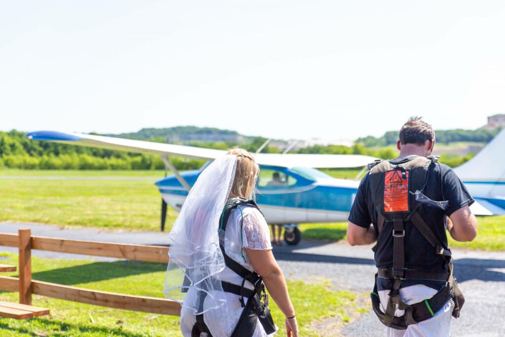 A bride and groom wearing parachute harnesses walk towards a small blue and white airplane on a sunny day. The bride is wearing a white veil. Grass and a wooden fence are visible in the background.