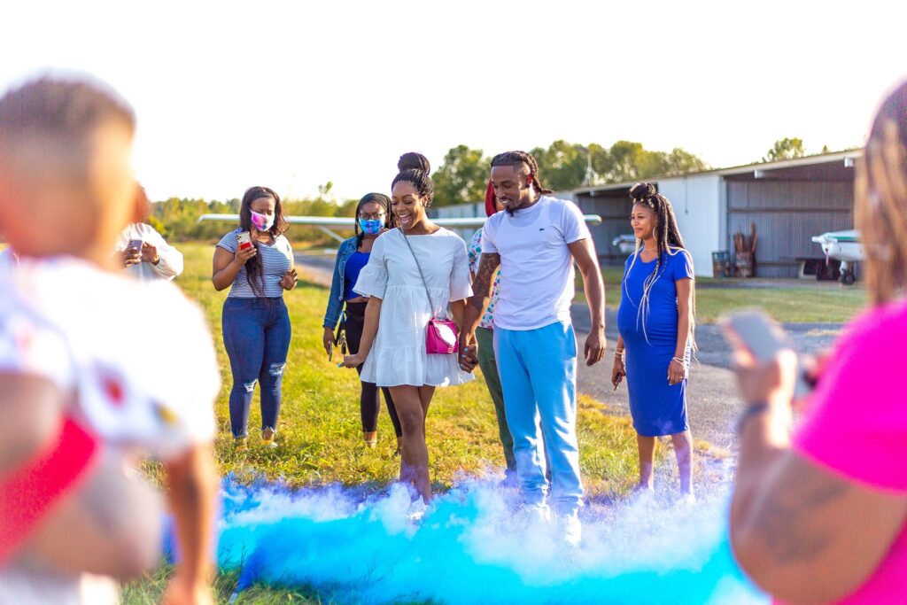 A joyful outdoor gender reveal event captures a couple walking hand in hand through blue smoke, surrounded by excited onlookers and photographers. The setting is sunny, with a grassy area and an airplane hangar in the background.