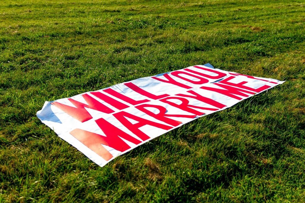 A large banner sprawled on green grass with bold red letters reads, "WILL YOU MARRY ME?" The edges of the banner are slightly uneven, suggesting it was quickly laid out for a proposal.