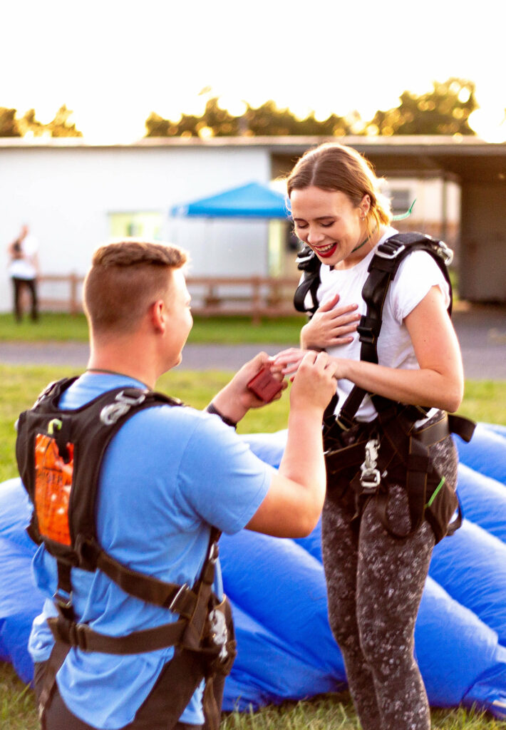A man kneels and presents a ring to a surprised woman, both wearing skydiving gear. They are outdoors, with a blue parachute laid on the grass. The woman holds her chest, smiling widely. A building and trees are in the blurred background.