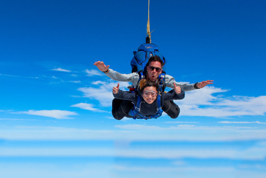 Two people are skydiving tandem style against a clear blue sky with scattered clouds. The instructor at the back and the participant in front both have their arms outstretched. The participant is giving a thumbs-up and smiling.