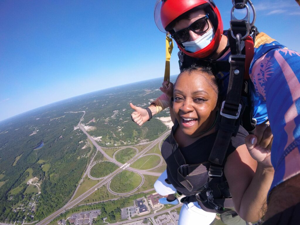 A woman and an instructor are tandem skydiving, giving a thumbs up and smiling at the camera. Below them, a sprawling landscape with roads and greenery is visible. The instructor wears a red helmet and goggles against a clear blue sky.