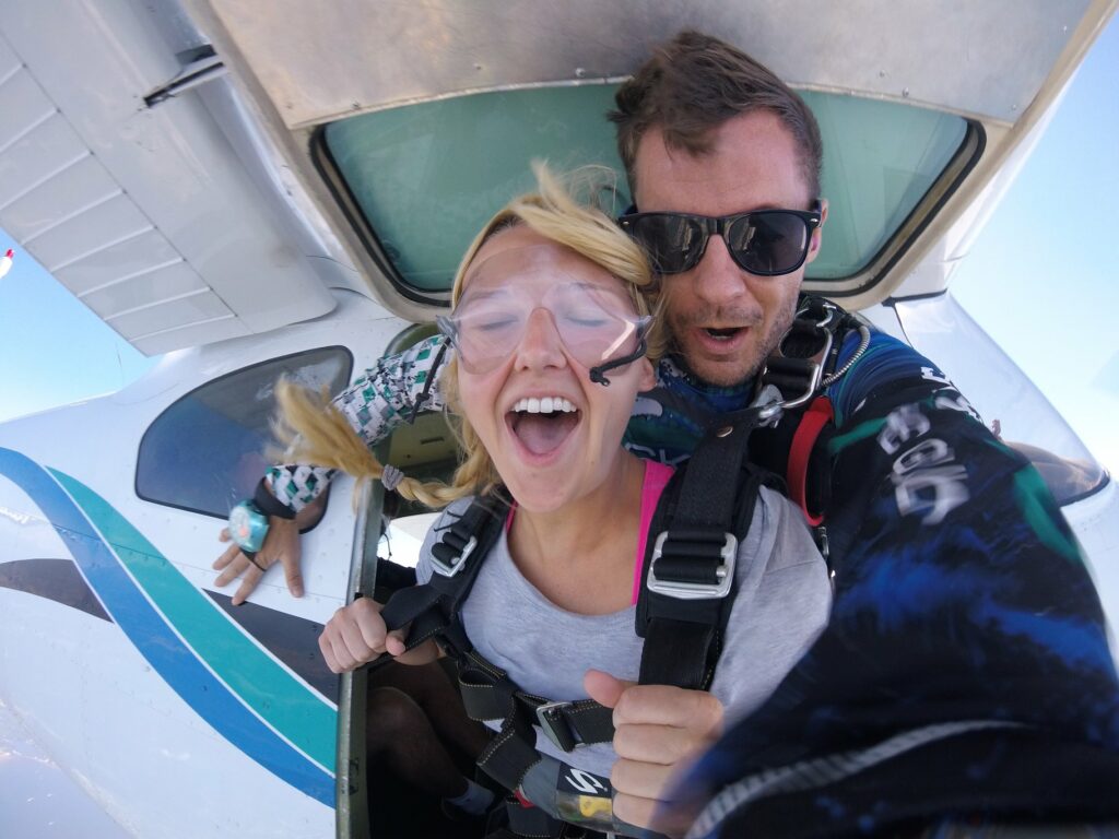 A woman and an instructor are skydiving tandem-style, just exiting an airplane. The woman is smiling widely, wearing goggles and a harness. The instructor, also in a harness, is behind her, wearing sunglasses. The sky is clear and blue.