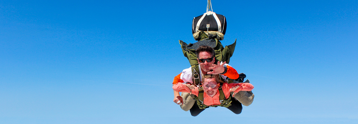 Two people tandem skydiving against a clear blue sky. The instructor, wearing sunglasses, is positioned above the other person, who is smiling with arms spread wide. Both are secured by a harness, and a parachute is visible at the top.