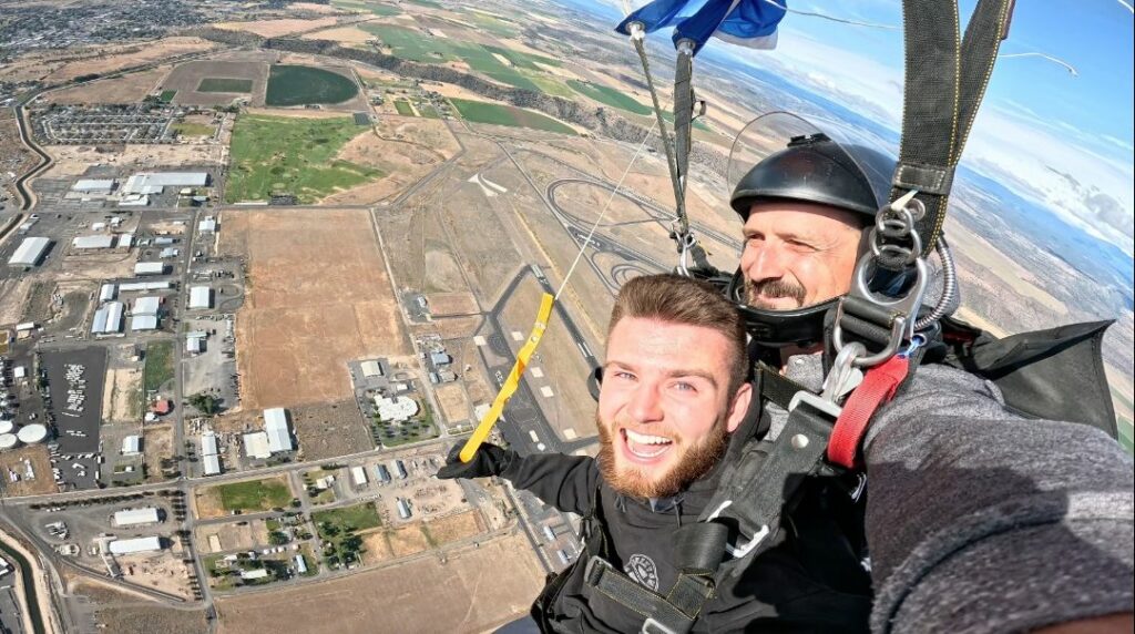 two male tandem skydivers smile for a selfie while flying under the parachute